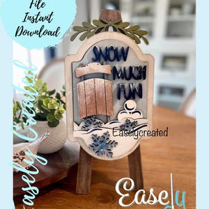 May include: A wooden sign with a white background and blue lettering that reads "Snow Much Fun". The sign features a wooden sled and a snowman. The sign is on a small wooden easel. The sign is a digital cut file.
