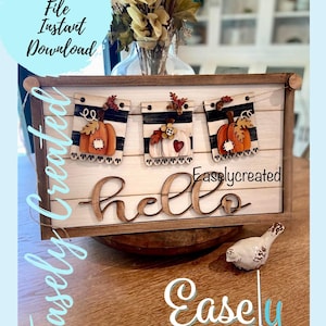 May include: A wooden sign with a black and white striped banner featuring pumpkins and fall leaves. The sign says "hello" in a decorative font and is framed in wood. The sign is sitting on a wooden surface with a small white bird figurine.