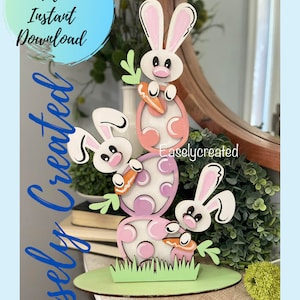 May include: A digital cut file for a three-tiered Easter bunny and egg decoration. The bunnies are holding carrots and the eggs are decorated with polka dots. The base is green and has a grass design. The text "Easlycreated" is visible on the image.