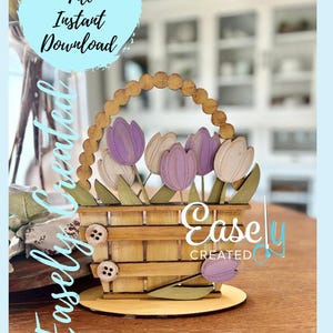 May include: A wooden basket with a purple and white tulip flower arrangement. The basket has a brown wooden base and a light brown wooden handle. The basket is decorated with buttons and the text "Easley Created".