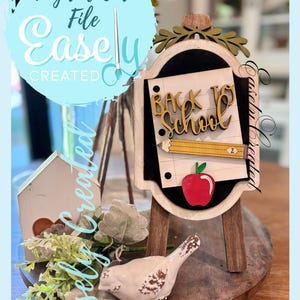 May include: A wooden easel with a chalkboard, featuring a paper with "Back to School" in gold lettering, a pencil, and a red apple. The easel is on a wooden base, with a small bird and house nearby.