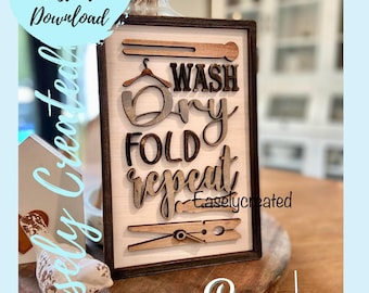 May include: Wooden framed sign with the words "WASH DRY FOLD REPEAT" in a mix of fonts. The sign includes wooden cutouts of a hanger, clothespin, and other laundry-related elements. The sign is on a wooden surface.