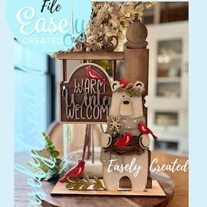 May include: Wooden winter-themed decoration featuring a bear with a sign that reads "Warm Winter Welcome." The bear is adorned with a wreath and surrounded by red cardinal figures. The words "Digital Cut File" and "Easely Created" are also visible.