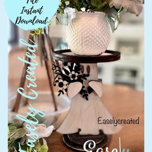 May include: A white wooden ghost figurine with black floral accents, wearing a white ceramic pot with a textured surface as a hat. The ghost is standing on a tiered wooden stand with a black metal base. The text "Easlycreated" is visible on the stand.