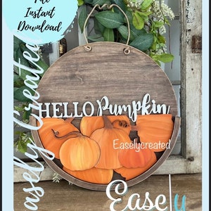Pu&ograve; includere: Un cartello rotondo in legno con un manico in corda, che presenta il testo "HELLO PUMPKIN" e un semicerchio di zucche arancioni. Il cartello &egrave; decorato con un motivo a venature di legno marrone e il testo "Easlycreated" &egrave; visibile nell'angolo in basso a destra.