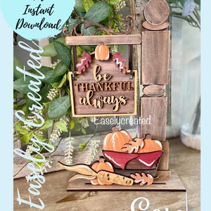 May include: A wooden sign with the text "be thankful always" hanging from a wooden post with a basket of pumpkins and corn on the bottom. The sign is decorated with fall leaves and the post is decorated with a wooden ball.