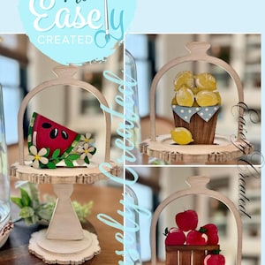 May include: Wooden decorative stands with fruit-themed cutouts. One stand features a watermelon slice with flowers, another has lemons in a basket, and a third displays red apples in a wooden crate. The stands have a light wood finish.