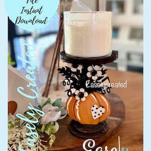 May include: A wooden pumpkin candle holder with a white candle, black and white flowers, and a black heart with white polka dots. The holder is on a wooden surface with green foliage.