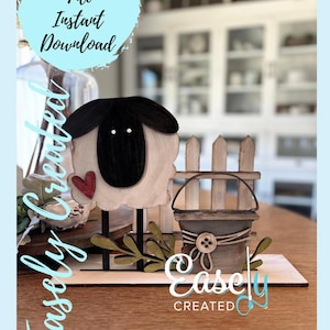 May include: A digital cut file for a wooden craft project featuring a sheep, a fence, and a bucket. The sheep is white with a black face and a red heart. The fence is white and the bucket is brown with a white button. The text "Easily Created" is on the base.