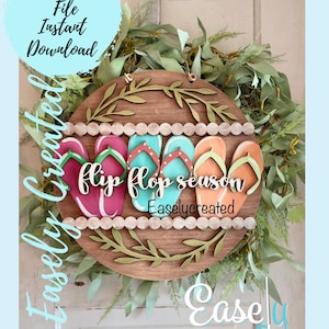 May include: A wooden round sign with a wreath of green leaves around the edge. The sign says "flip flop season" with a row of colorful flip flops on each side of the text. The sign is hanging on a white wooden door.