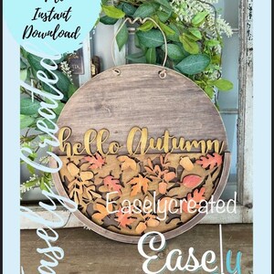 Pu&ograve; includere: Un cartello rotondo in legno con il testo "hello autumn" in lettere dorate. Il cartello &egrave; decorato con una cornice in legno marrone e un design ritagliato di foglie autunnali in arancio, rosso e giallo.