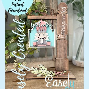 May include: A wooden sign featuring a nutcracker design and the word "Welcome". The sign is attached to a wooden post with a green leafy sprig at the base. The sign is labelled "Easly Created".