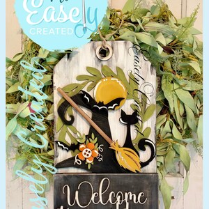 May include: A wooden Halloween-themed sign with a black witch's hat, broom, and cat silhouette. The sign includes the text "Welcome to our home" and "Easely Created". The background features a wreath of green leaves.