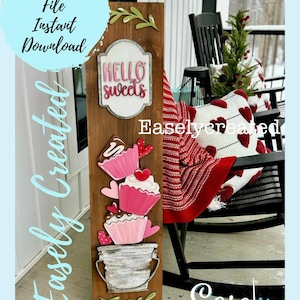 May include: A wooden sign with the text "Hello Sweets" and a stack of three pink and white cupcakes in a galvanized bucket. The sign is decorated with green leaves and is leaning against a black rocking chair on a porch.