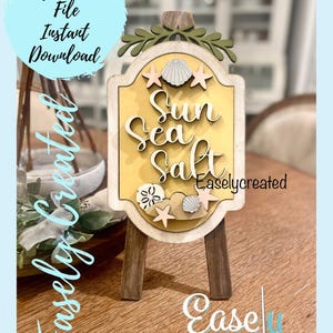 May include: A wooden sign with a yellow background and the text "Sun Sea Salt" in a decorative font. The sign is decorated with seashells, starfish, and a small sand dollar. The sign is on a small wooden easel.