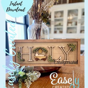 May include: A wooden sign with the word "HOLY" in large, decorative letters. The letters are surrounded by greenery and a small manger scene with a star on top. The sign is framed in a brown wood frame. The sign is sitting on a wooden table with a vase of dried flowers in the background.