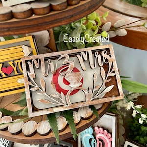 May include: Wooden tiered tray with various signs. The top tier has a sign that says "Mr. & Mrs." with a ring and floral accents. The middle tier has a sign that says "You're the best" with a pencil and heart. The bottom tier has signs that say "Grad", "Son", and "Class of".