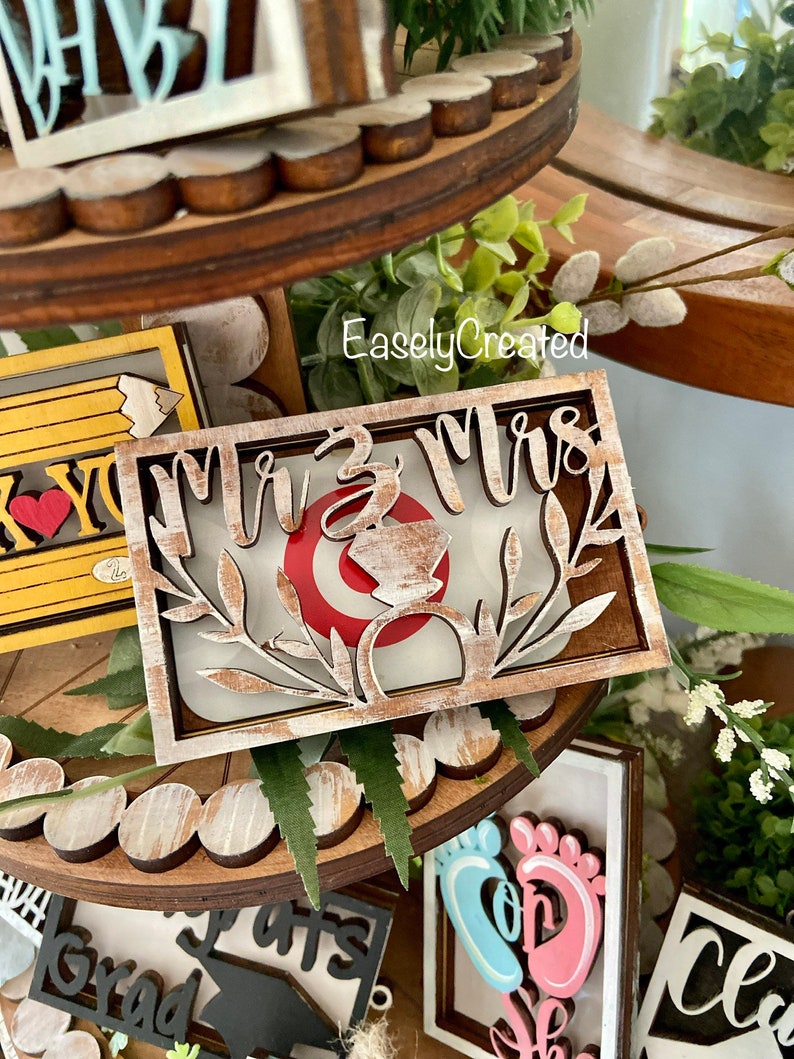May include: Wooden tiered tray with various signs. The top tier has a sign that says "Mr. & Mrs." with a ring and floral accents. The middle tier has a sign that says "You're the best" with a pencil and heart. The bottom tier has signs that say "Grad", "Son", and "Class of".