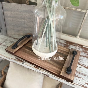 May include: A wooden tray with black metal handles. The tray is stained brown and has a white painted design. The tray is sitting on a white painted table with a vase of dried flowers.