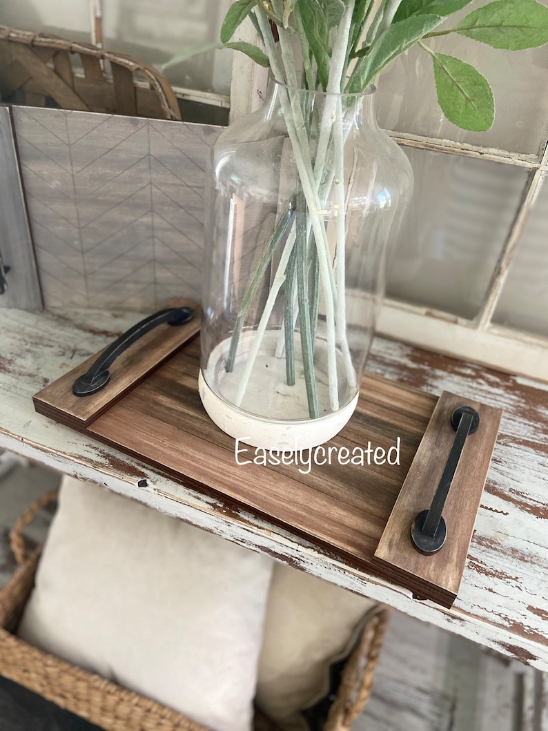 May include: A wooden tray with black metal handles. The tray is stained brown and has a white painted design. The tray is sitting on a white painted table with a vase of dried flowers.