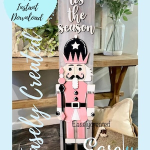 May include: A pink and black nutcracker with a white star on its hat, standing in front of a wooden sign that says "tis the season".