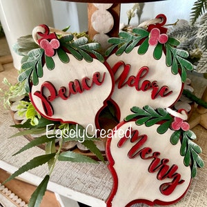 May include: Three wooden Christmas ornaments with red and green painted details. The ornaments are shaped like teardrops and have the words "Peace", "Adore", and "Merry" written on them in red.