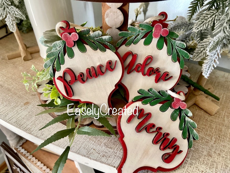 May include: Three wooden Christmas ornaments with red and green painted details. The ornaments are shaped like teardrops and have the words "Peace", "Adore", and "Merry" written on them in red.