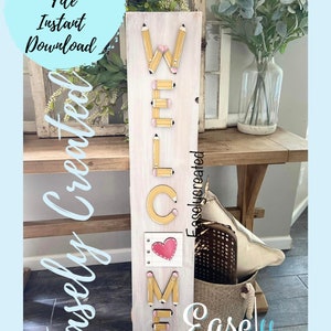 May include: A white wooden sign with the word "WELCOME" spelled out using yellow pencils. The sign has a pink heart with a white outline in the centre of the word "COME". The sign is leaning against a wooden table with a wicker basket and a white pillow.