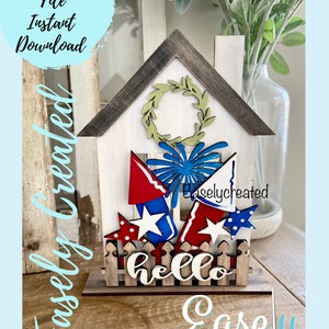 May include: A wooden house-shaped sign with a red, white, and blue patriotic theme. The sign features fireworks, stars, and the word "hello" on a picket fence.