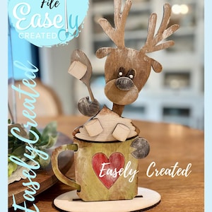 May include: A wooden reindeer-themed decoration with a mug design. The mug has a red heart and the text "Easely Created". The reindeer has antlers and is holding a spoon with marshmallows. The words "Digital Cut File" are in a blue circle.