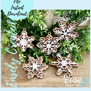 Puede incluir: Cinco adornos de copo de nieve de madera con un diseño pintado en blanco. Los copos de nieve están dispuestos sobre una superficie de madera con follaje verde en el fondo. El texto "Easly Created" es visible en la esquina inferior derecha de la imagen.
