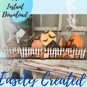 May include: A Halloween-themed wooden craft project featuring a row of houses, a moon with bats, a black cat, and pumpkins. The houses have numbers on them, and there is a white picket fence in front of them. The project is on a whitewashed wooden surface. The text "Easley Created" is visible in the image.