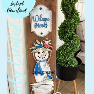 May include: A wooden sign with a snowman, a welcome friends sign, and a green bush in a black pot. The sign is decorated with blue and red accents. The text "Easlycreated" is visible on the sign.