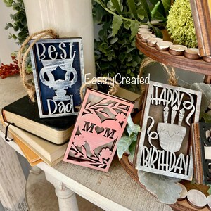 May include: Three wooden signs with white painted lettering. The first sign says "Best Dad" in blue and white. The second sign says "Mom" in pink with a heart. The third sign says "Happy Birthday" in grey and white.