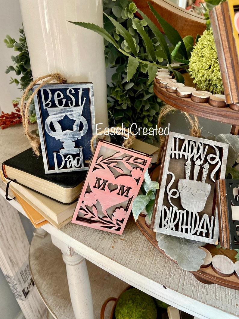 May include: Three wooden signs with white painted lettering. The first sign says "Best Dad" in blue and white. The second sign says "Mom" in pink with a heart. The third sign says "Happy Birthday" in grey and white.