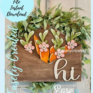May include: A wooden envelope with a "hi" sign and three carrots with green leaves and pink flowers. The envelope is surrounded by green leaves.