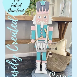 May include: A wooden nutcracker figurine painted in shades of pink, blue, and gray. The nutcracker is holding drumsticks and a drum with a star on it. The text "Easlycreated" is printed on the drum. The nutcracker is standing on a gray base.
