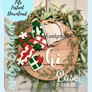 May include: A wooden round sign with the word "hi" in white script, surrounded by green leaves. There are four red and green gift boxes and a white bucket with a handle on the sign. The sign is surrounded by a wreath of green leaves.