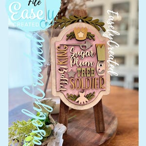 May include: A wooden sign with a pink and green design, featuring text related to the Nutcracker ballet. The sign is mounted on a small wooden easel. The words "Sugar Plum Tree Soldier" are visible.