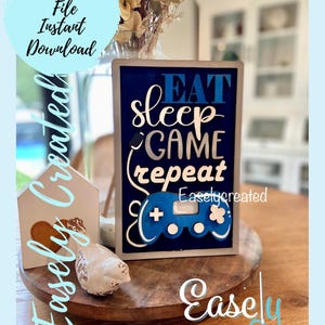 Könnte beinhalten: Ein marineblaues Holzschild mit den Worten "EAT SLEEP GAME REPEAT" in Weiß und Blau. Unter dem Text ist ein blauer Gamecontroller abgebildet. Das Schild steht auf einem Holztablett mit dekorativen Elementen.