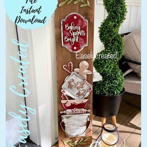 Op de afbeelding: Een houten bord met een rood en wit bord waarop "Baking Spirits Bright" staat en een peperkoekman, een deegroller, een kom en een bakplaat met koekjes erop. Het bord is versierd met groene bladeren en rode en witte pepermuntstokken.