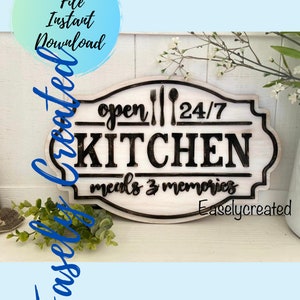 May include: A white wooden sign with black lettering that reads "open 24/7 KITCHEN meals & memories". The sign has a decorative border and a cutout of a fork, knife, and spoon. The sign is on a white surface with a green plant in the background.
