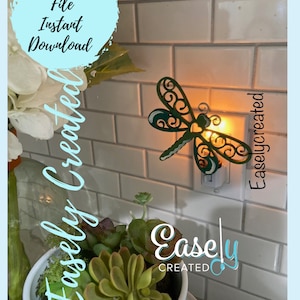 May include: A green dragonfly nightlight plugged into an electrical outlet. The dragonfly has intricate details and is illuminated by a warm light. The text "Easley Created" is visible on the wall.