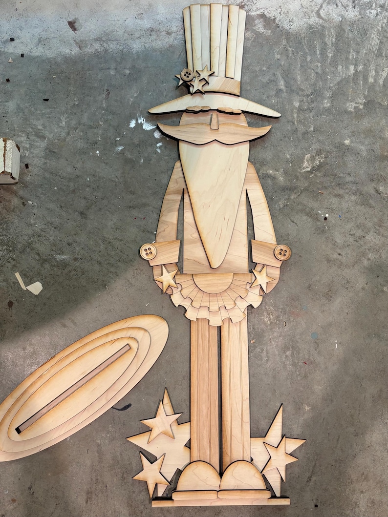 May include: Wooden cutout of a patriotic Uncle Sam figure with a tall hat, stars, and a beard. The figure is holding a banner with stars and is standing on a base with stars. The figure is made of unfinished wood.