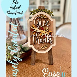 May include: A wooden sign with the text "Give Thanks" in a decorative font. The sign is decorated with fall leaves and is on a small easel.