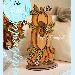 May include: A wooden pumpkin stack decoration with three pumpkins, leaves, and flowers. The pumpkins are orange and brown, with green leaves and small flower accents. The text "Easely Created" is visible.