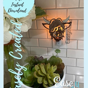 Puede incluir: Una luz nocturna de madera con forma de vaca de las tierras altas conectada a una toma de corriente. La vaca es marrón con la cara blanca y tiene una luz que brilla a través de ella. La luz nocturna está en una pared de azulejos blancos.