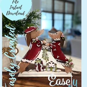 May include: Two wooden dachshund figurines wearing red and white Christmas outfits, holding a sprig of holly and standing on a snowy base with presents. The figurines are on a wooden surface with the text "Easley Created" in white.