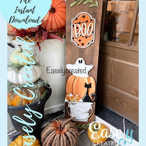 Puede incluir: Un letrero de madera con un fantasma, una calabaza y un gato negro. El letrero también tiene un letrero "BOO" separado con lunares naranja y negro. El letrero está apoyado contra una puerta con una corona de hojas de otoño.