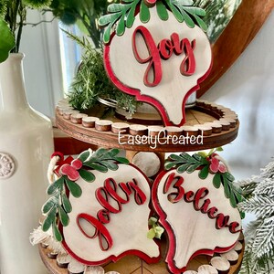 May include: Three wooden Christmas ornaments with red and green accents. The ornaments are shaped like teardrops and have the words "Joy", "Jolly", and "Believe" written on them in red.